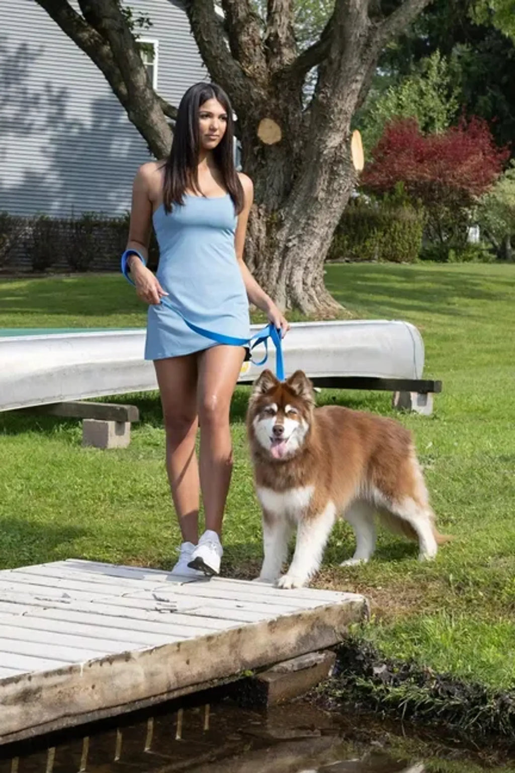 Woman in a blue dress standing with a dog on a wooden dock in a park-like setting.