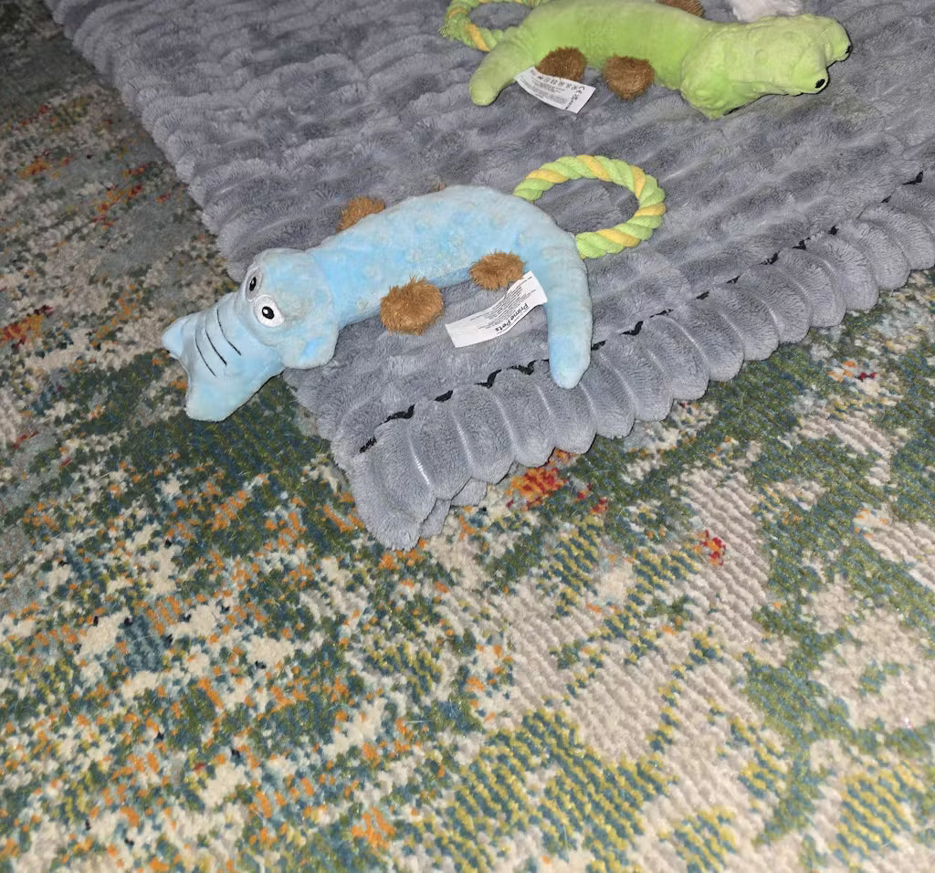 Plush toy alligator on a textured gray blanket with another plush toy in the background.